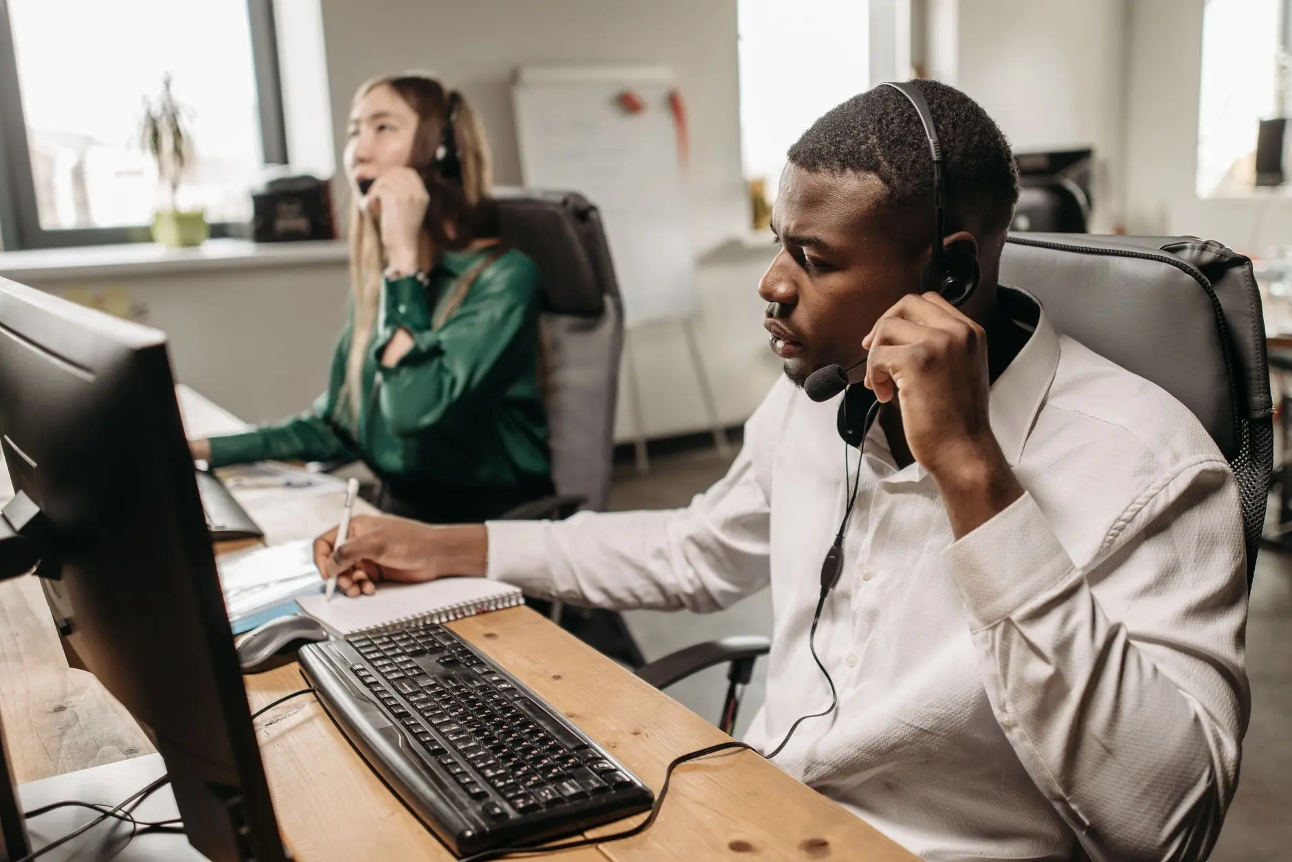 Two colleagues in a warm office: a man wearing a headset at a computer and a woman standing behind, illustrating collaborative AI receptionist setup.