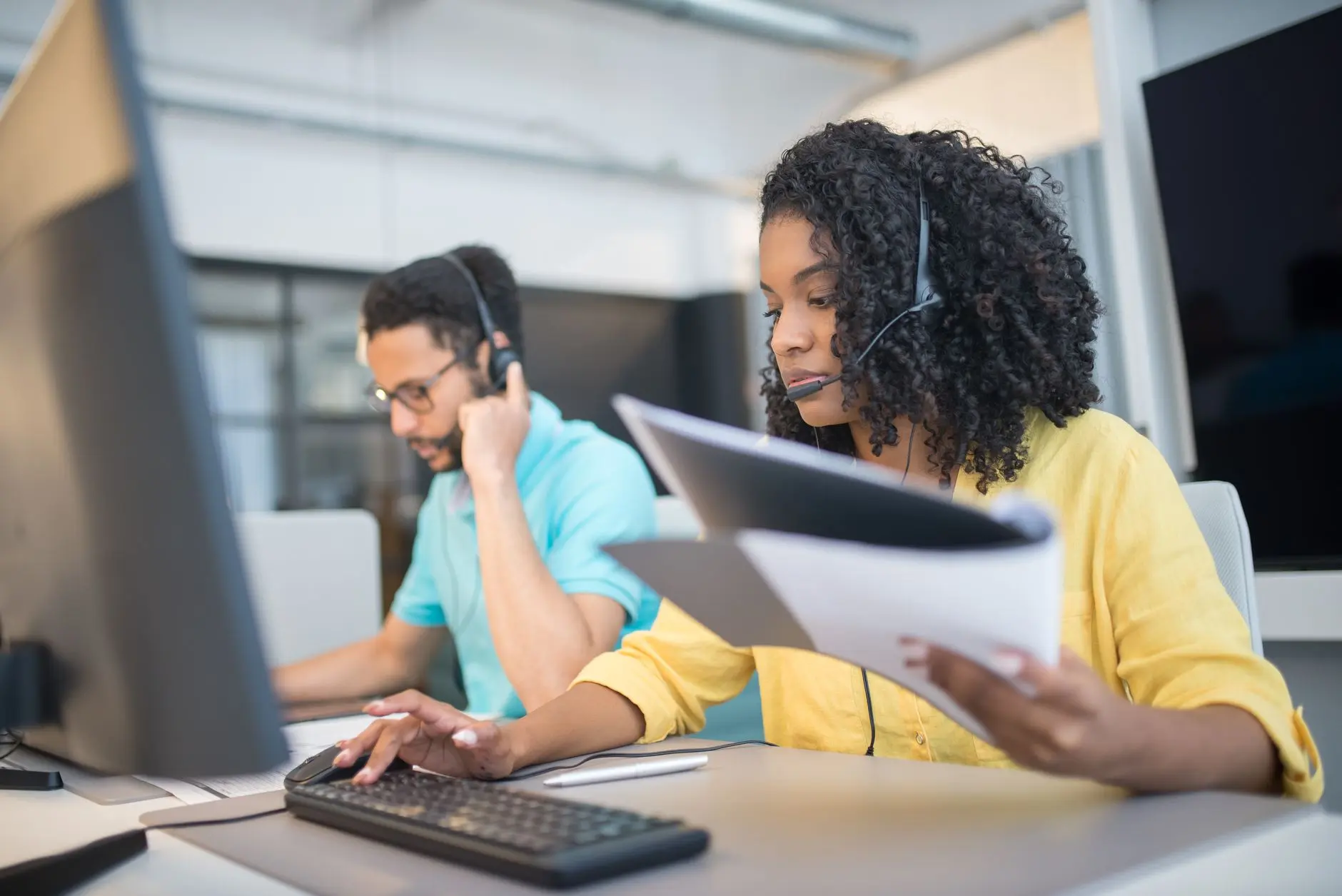 Two colleagues in a bright modern office: man with headset at a desk with multiple screens, woman reviewing data-heavy papers.