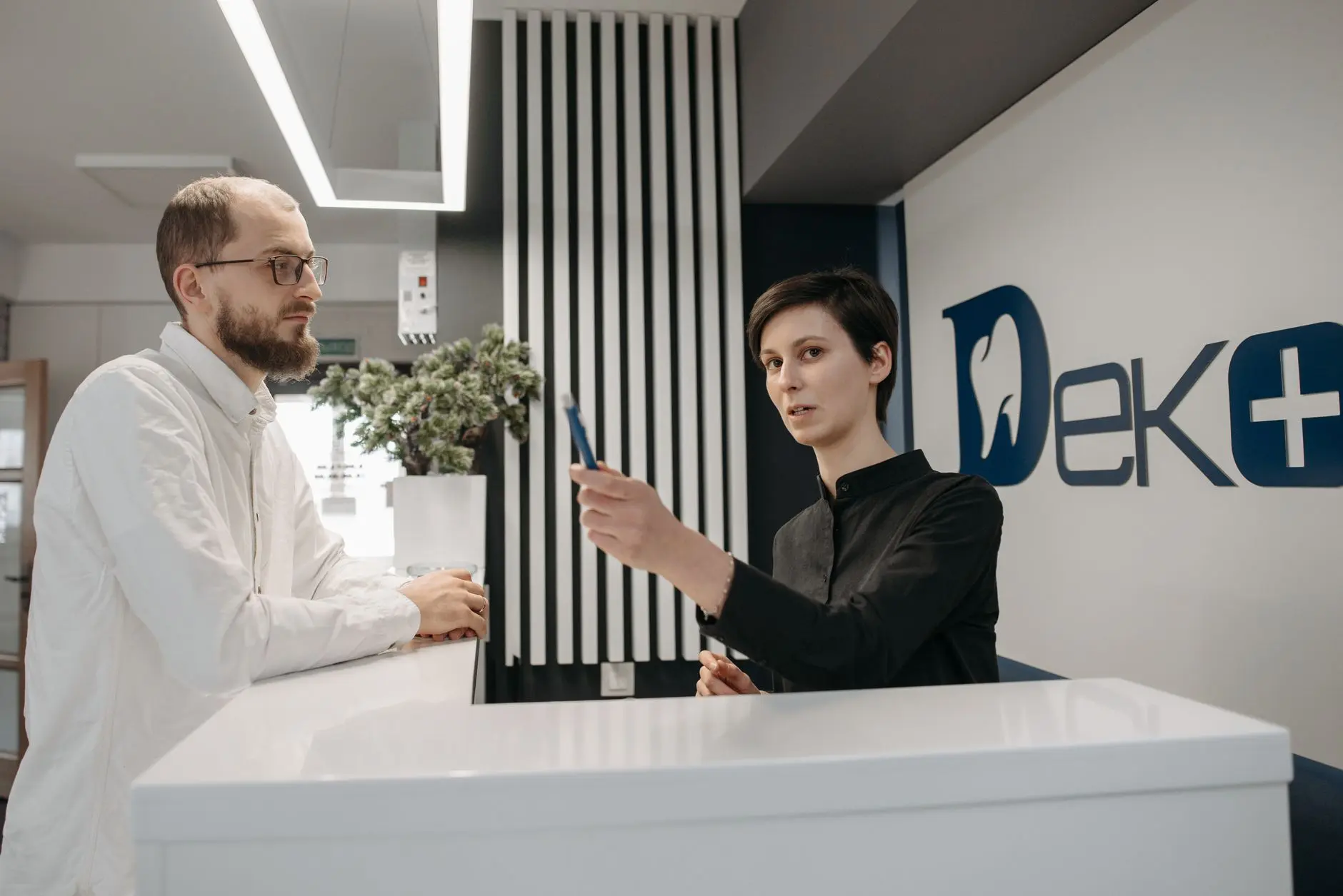 Two professionals at a modern reception desk; one holds a card while the other works at counter, with plants and DEKE sign.