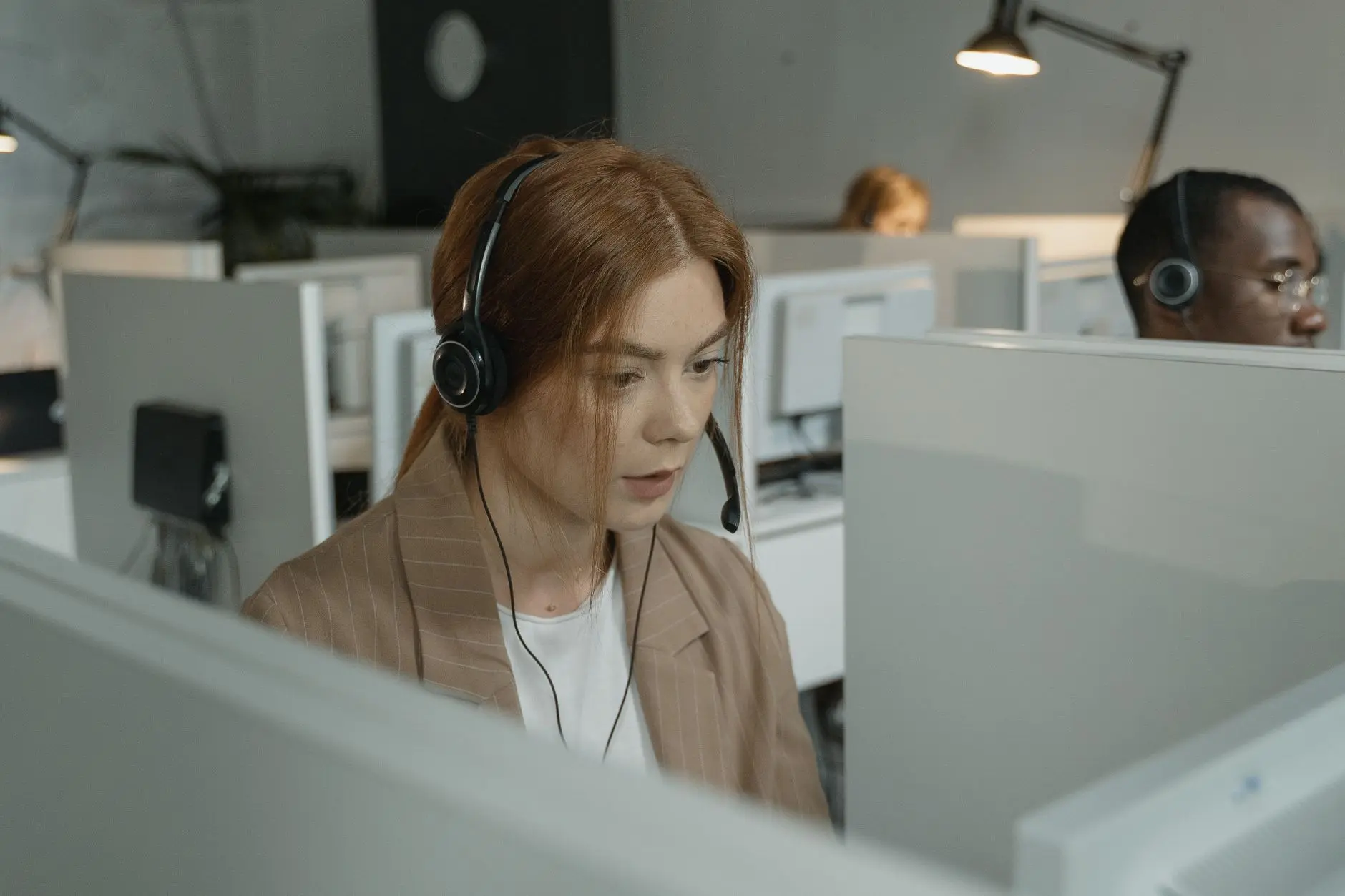 Focused woman in a blazer sits at a computer in a modern office, wearing headphones, while another person works at a nearby desk.