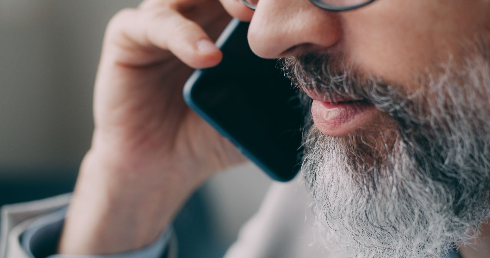 South Asian man with glasses and a beard on a phone call at a desk in an office; shirt reads 'Purpose'.