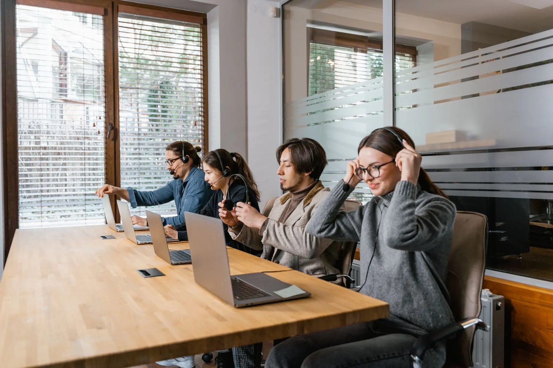 Four professionals sit around a wooden desk with laptops and headphones in a bright modern office with a whiteboard.