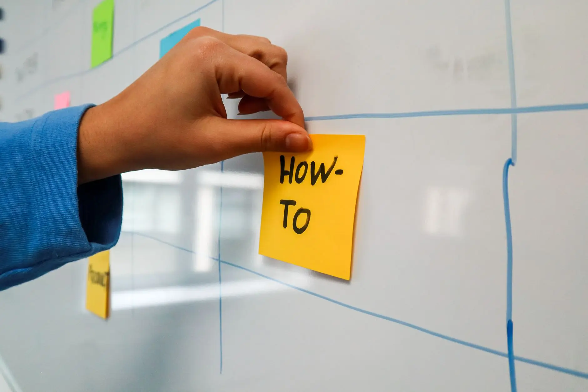 Office planning scene with a whiteboard, a hand placing a yellow Post-it labeled HOW TO, and a green checkmark.